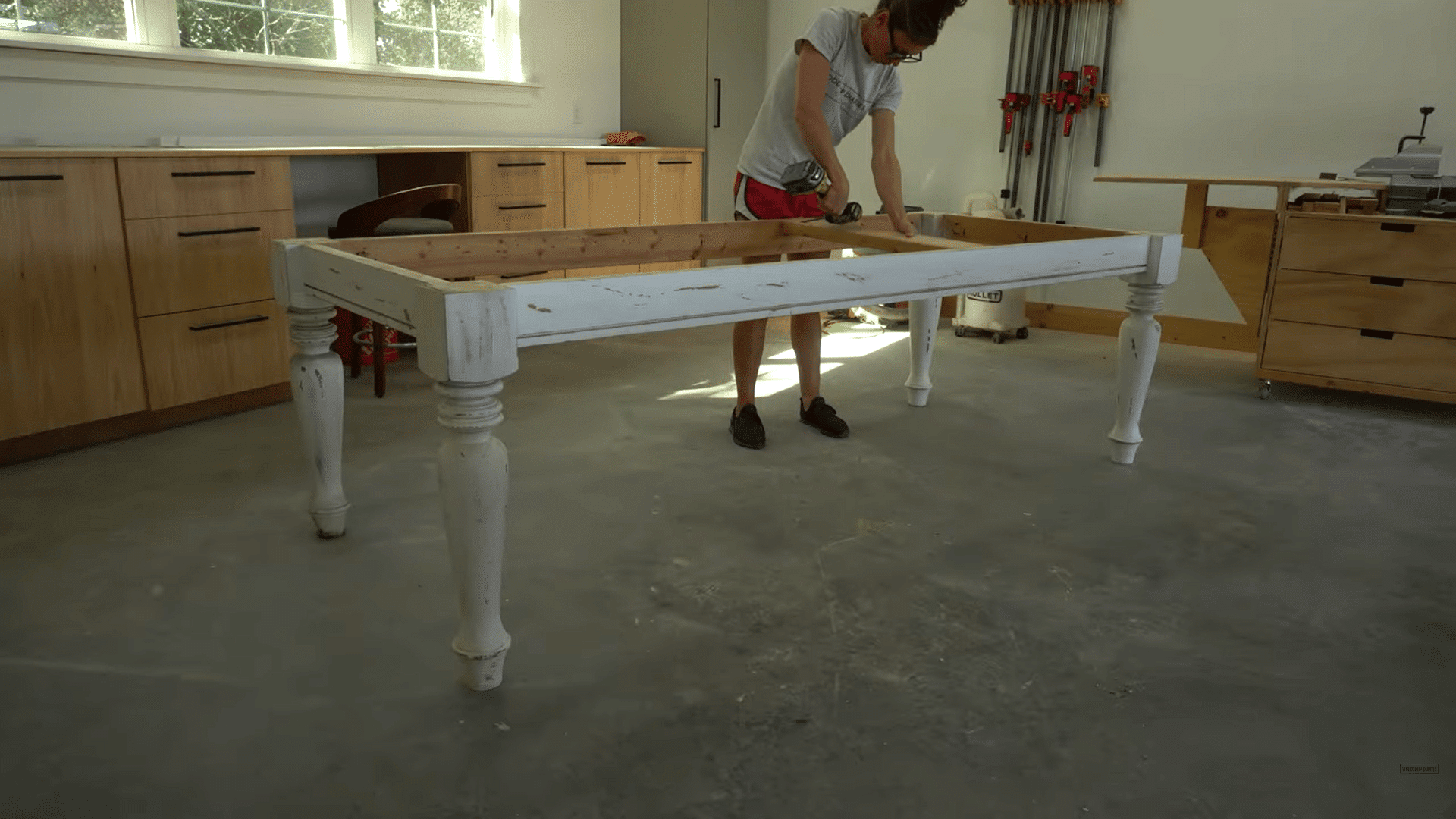 a person using a power drill to assemble a white wooden table frame inside a bright woodworking workshop