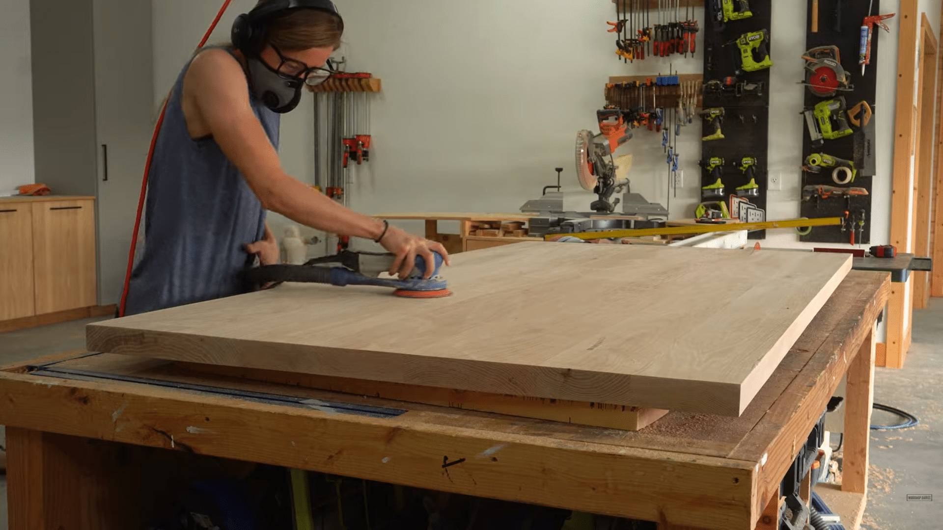 a person wearing safety gear sanding a large wooden tabletop inside a woodworking shop with tools on the wall