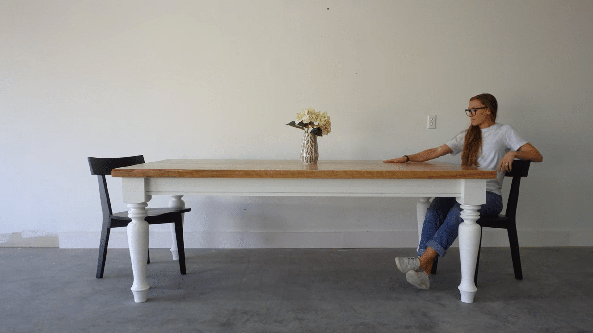 a woman sitting beside a completed farmhouse dining table showcasing the finished woodworking project in a simple room