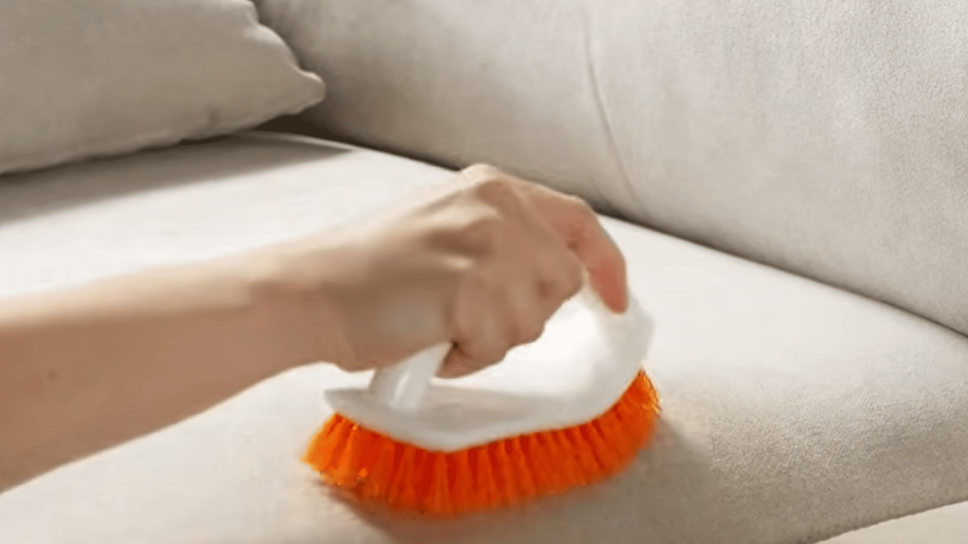 hand using an orange bristle brush to scrub and clean a light colored fabric couch cushion indoors