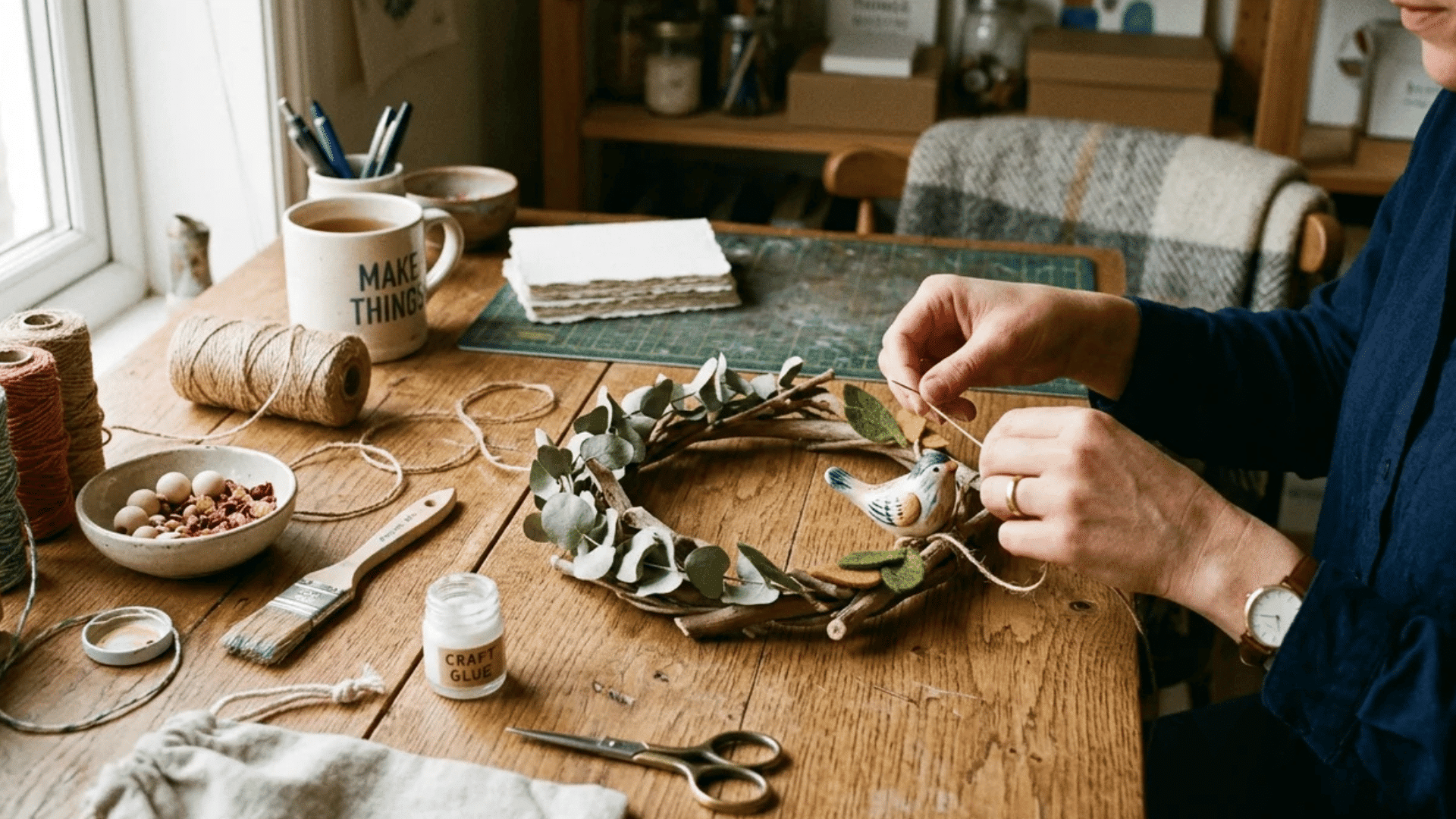 person crafting handmade wreath with twigs leaves and small bird decoration on wooden table with tools and materials nearby