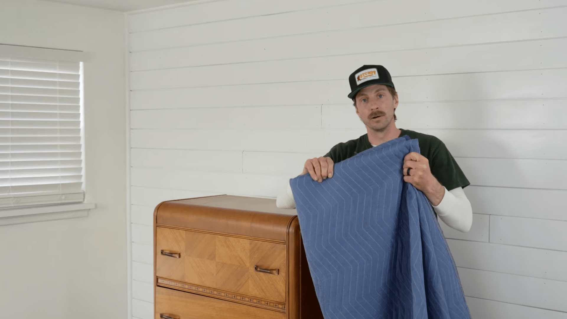 person holding a thick moving pad beside a wooden dresser to prepare furniture for safe moving