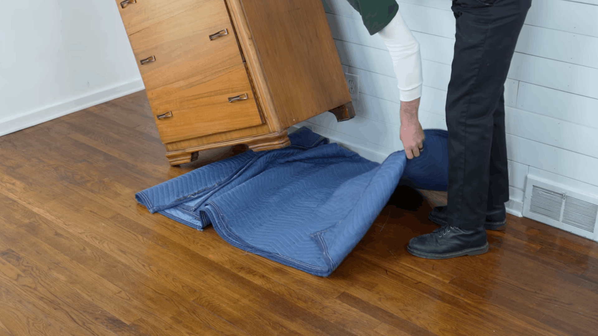 person placing a thick moving pad under a wooden dresser to protect floors during furniture moving