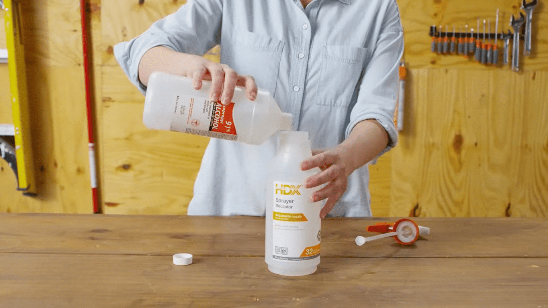 person pouring rubbing alcohol into a plastic spray bottle on a wooden workbench with tools hanging in background
