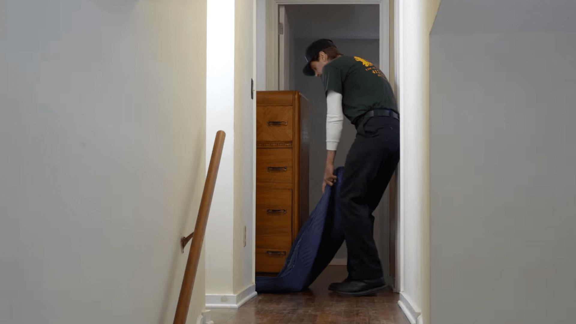 person pulling a wooden dresser on a moving pad through a narrow hallway while turning carefully around corners