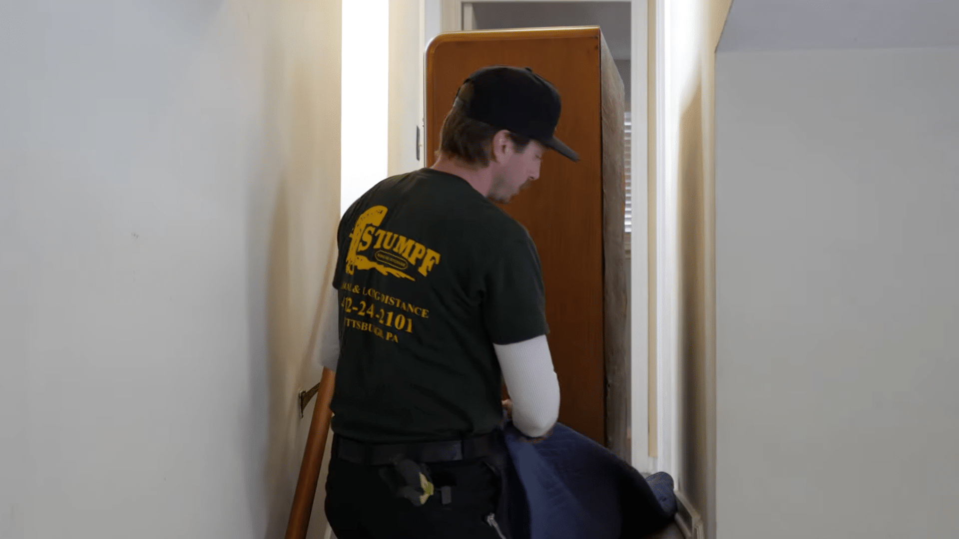 person rotating a wooden dresser in a narrow hallway while guiding it safely with a moving pad
