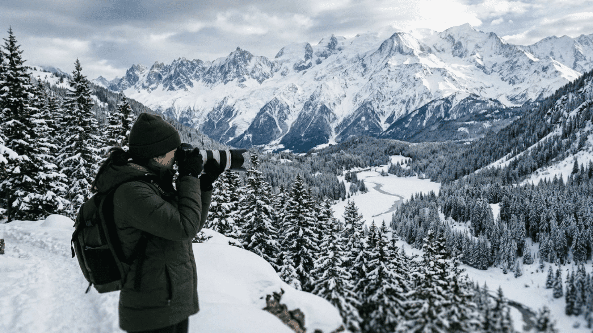 person taking photo with camera overlooking snowy mountain valley with trees and winding river under cloudy winter sky
