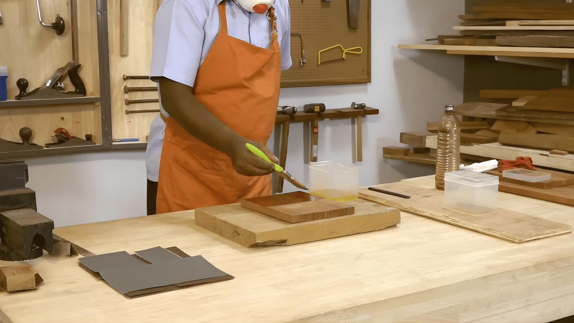person wearing apron applying liquid polish with brush onto wooden board in workshop showing how to polish wood furniture