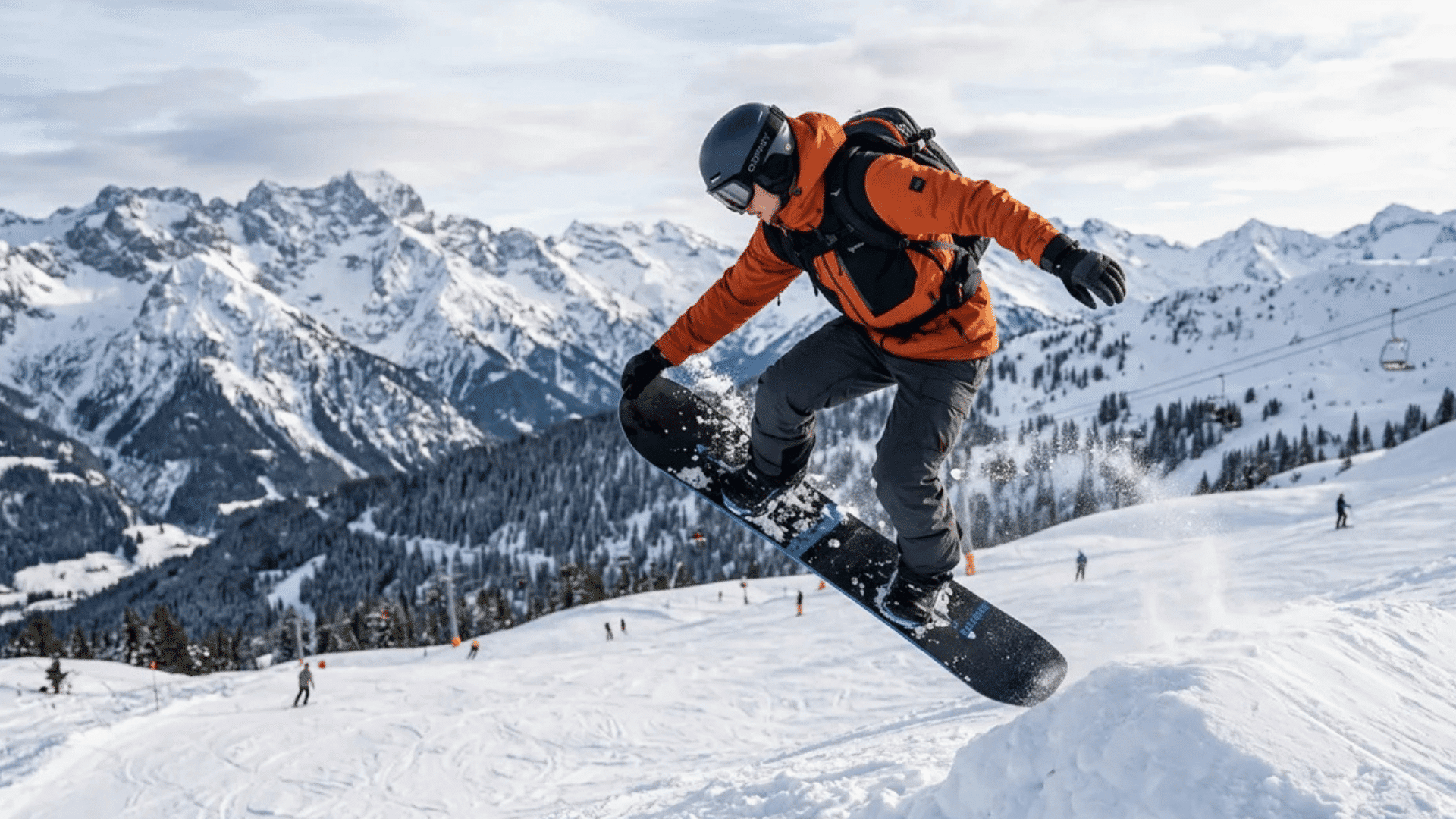 snowboarder in orange jacket performing jump on snowy slope with mountains and ski lifts in background under cloudy sky