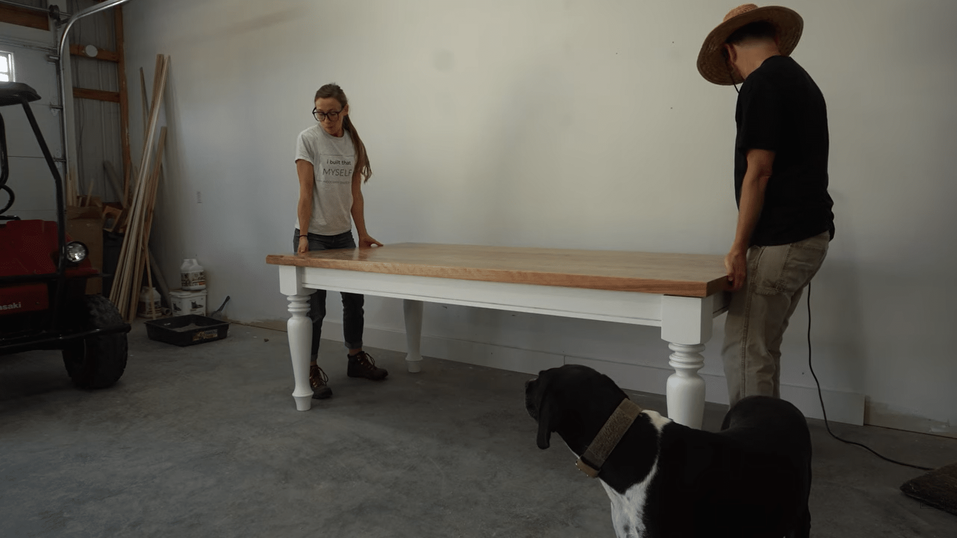 two people positioning a finished wooden farmhouse table in a garage workshop while a black dog watches nearby