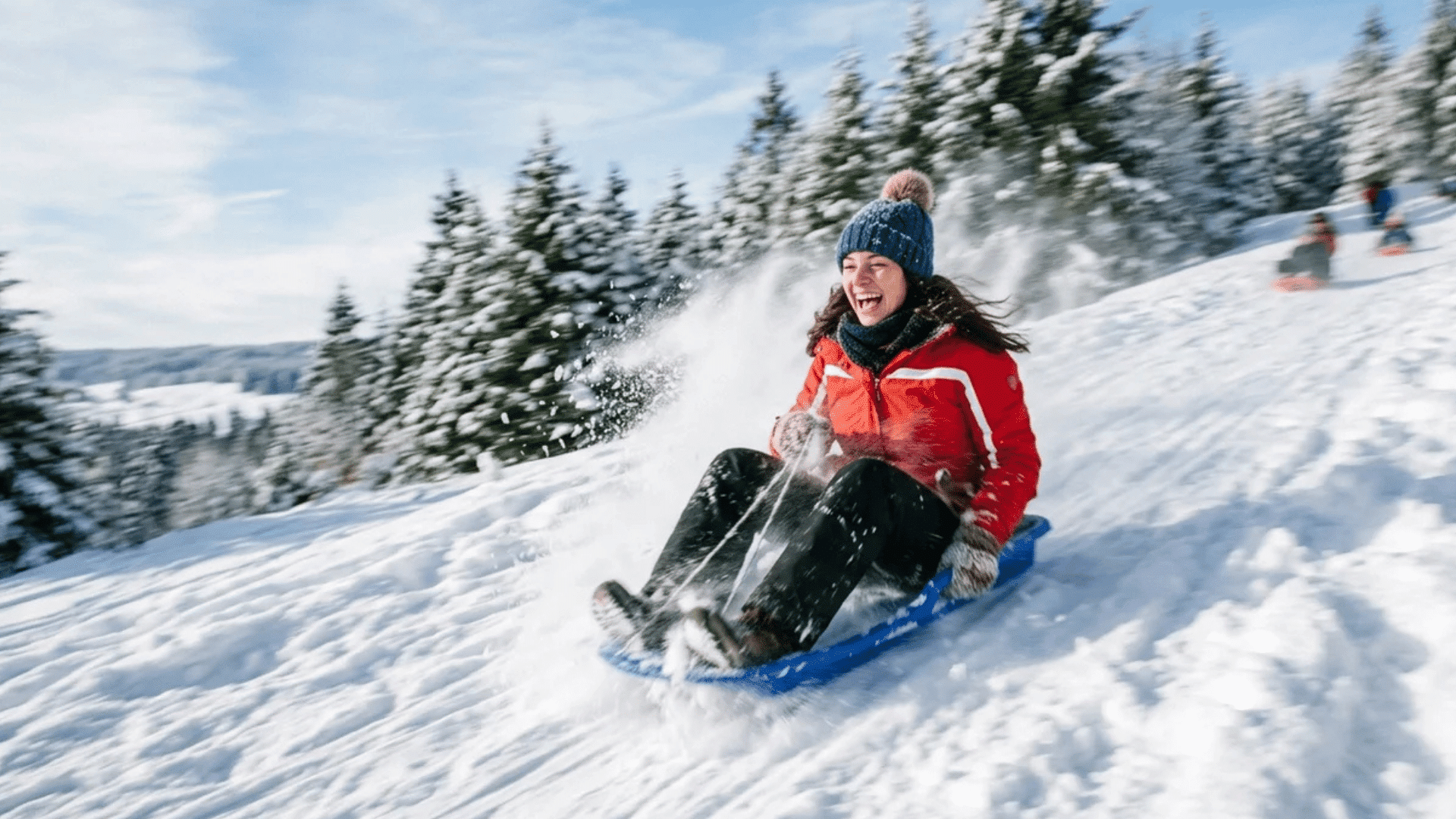 woman sledding down snowy hill wearing red jacket and blue hat with snow spraying around and pine trees in winter landscape