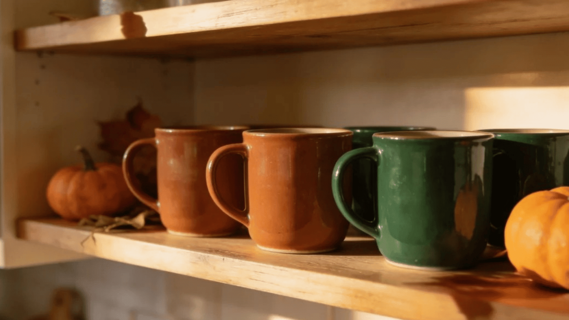 ceramic mugs on wooden kitchen shelves surrounded by pumpkins and autumn leaves in warm sunlight