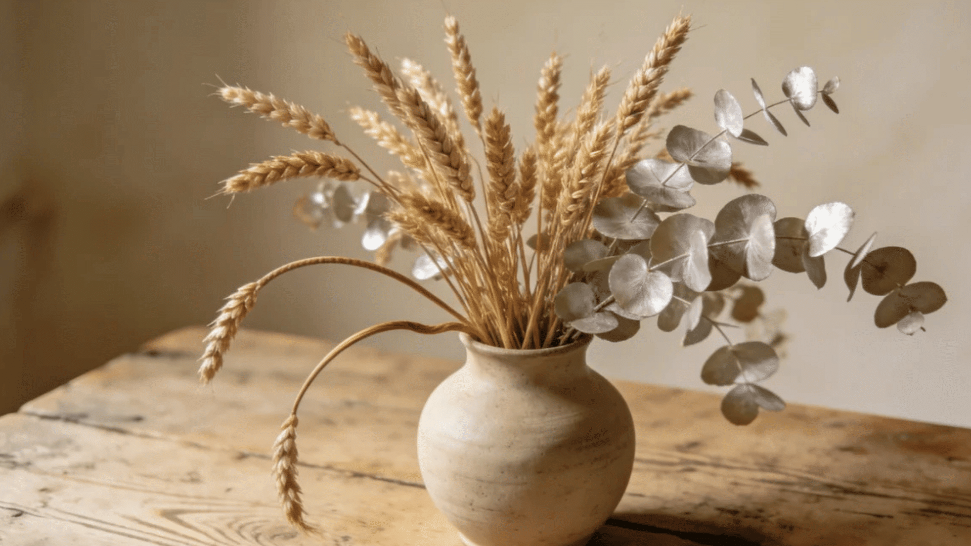 ceramic vase with dried wheat and eucalyptus stems placed on rustic wooden table in soft light