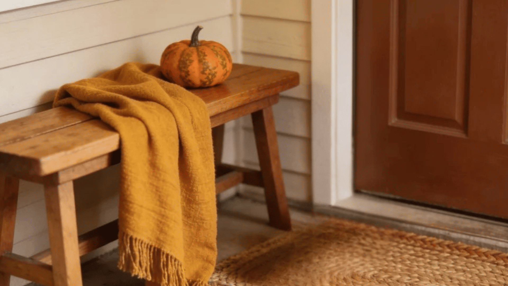 wooden porch bench with mustard blanket and pumpkins beside woven mat near front door
