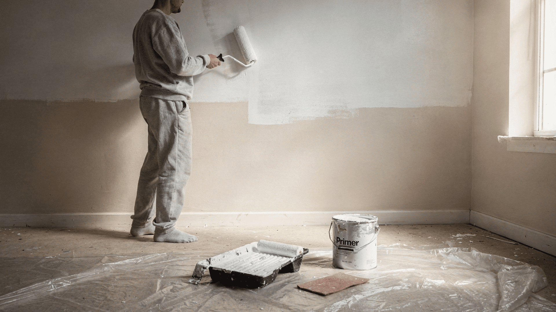 person rolling white paint onto wall during home renovation with primer can and sandpaper visible on drop cloth floor