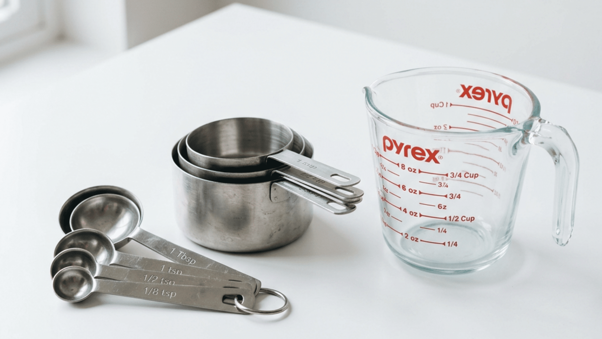 set of stainless steel measuring cups and measuring spoons and a glass measuring cup arranged on a clean white surface with soft natural light from a nearby window