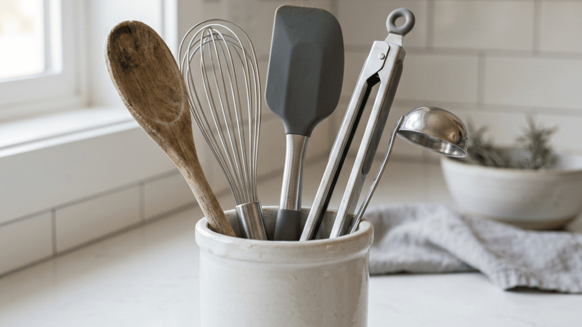 simple ceramic crock holding a wooden spoon and a whisk and a silicone spatula and tongs and a ladle on a white marble counter with soft natural light and a small plant in the background