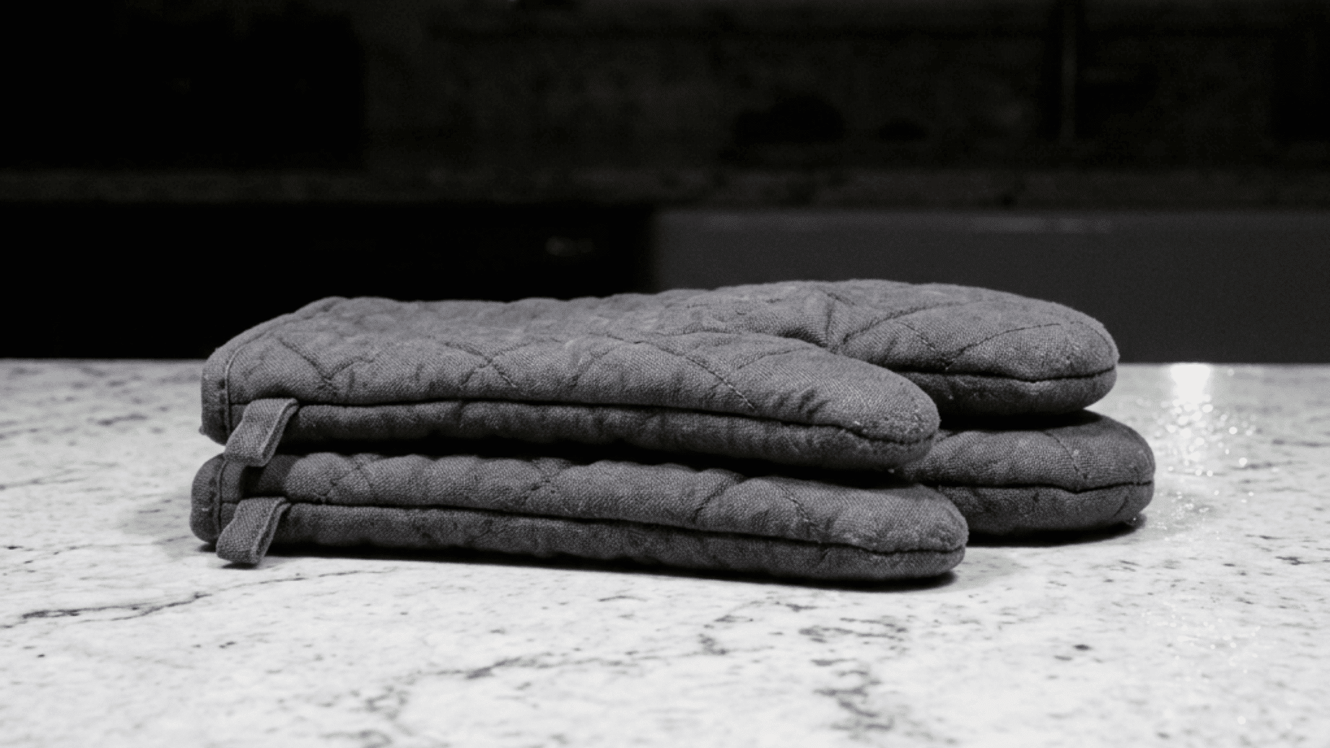 two charcoal grey quilted oven mitts folded and stacked neatly on a white marble counter with a softly lit background